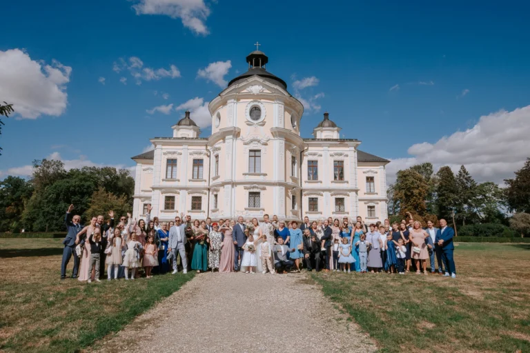 Skupinová fotografie novomanželů se svatebními hosty před kostelem.