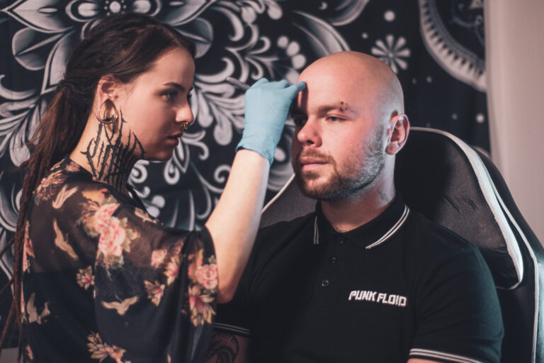 Potetovaná holka s dredama dělající piercing holohlavému muži na obočí