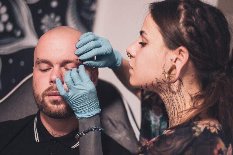 Potetovaná holka s dredama dělající piercing holohlavému muži na obočí