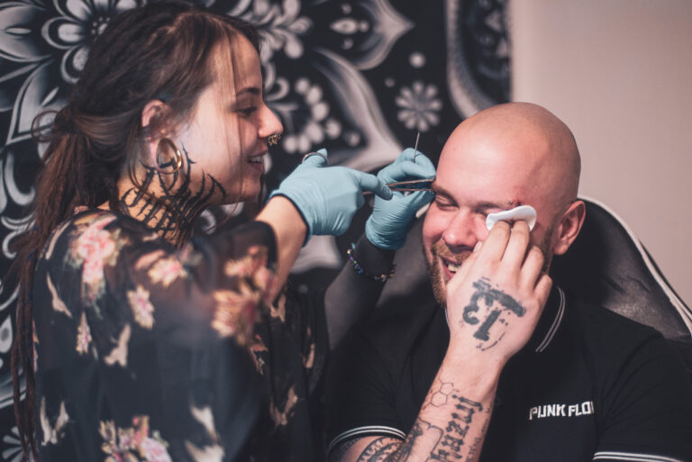 Potetovaná holka s dredama dělající piercing holohlavému muži na obočí