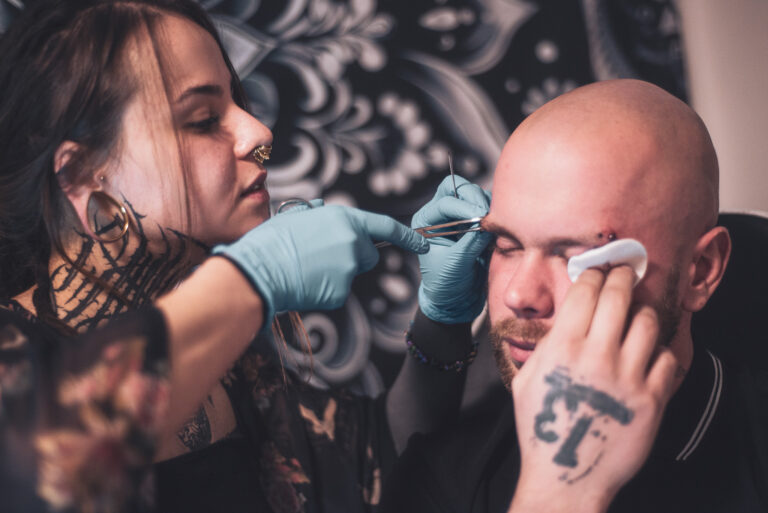 Potetovaná holka s dredama dělající piercing holohlavému muži na obočí