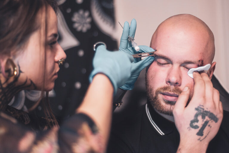 Potetovaná holka s dredama dělající piercing holohlavému muži na obočí