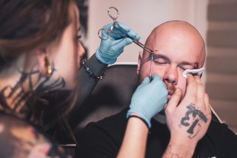 Potetovaná holka s dredama dělající piercing holohlavému muži na obočí