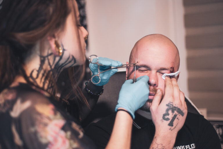 Potetovaná holka s dredama dělající piercing holohlavému muži na obočí